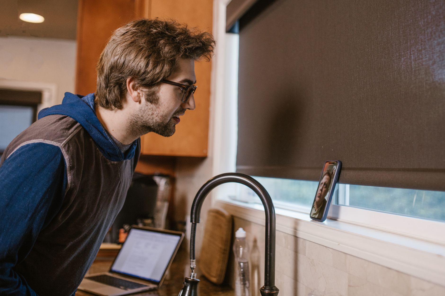 A man leans toward a phone propped by a kitchen window during a video call